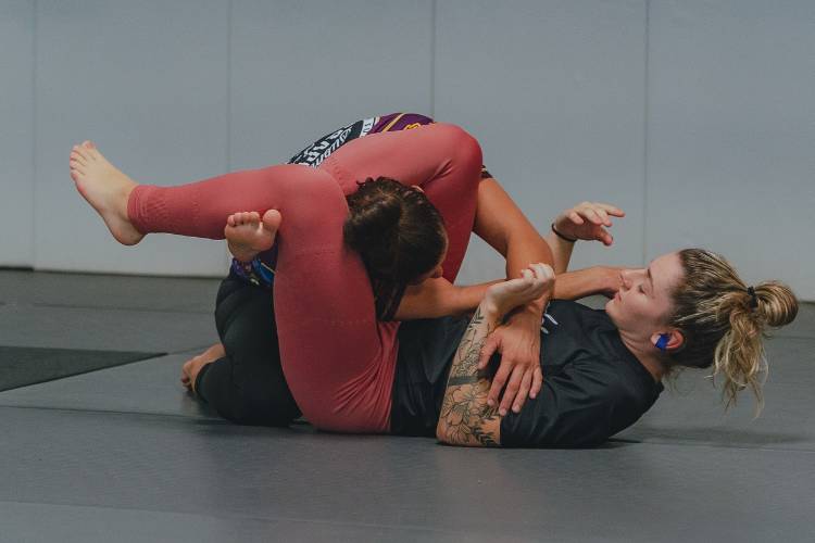 Two women practicing Brazilian jiu-jitsu on the mat, with one applying a leg-based control position during training.