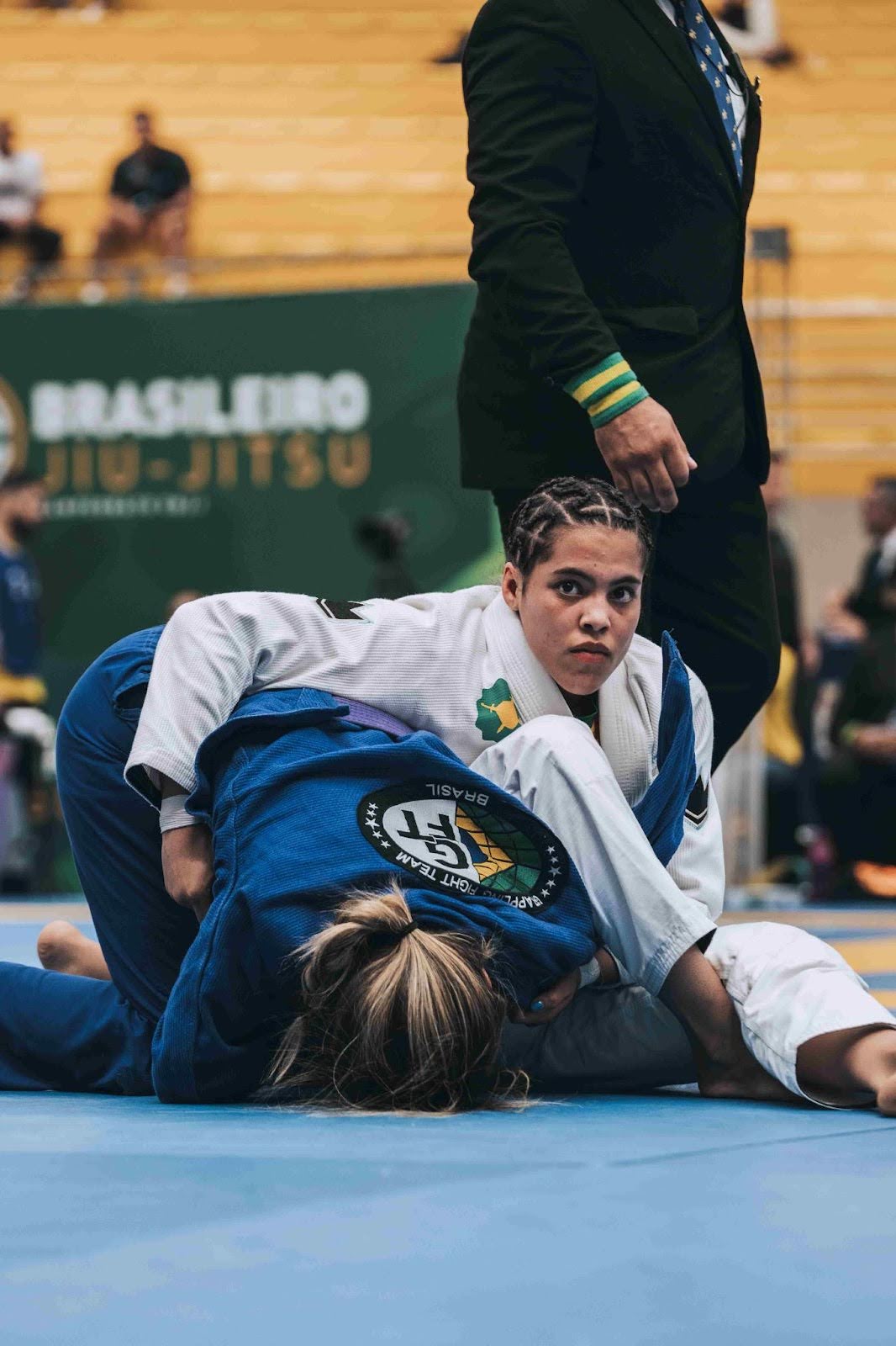 Two athletes competing in a Brazilian Jiu-Jitsu tournament, one controlling the other on the mat while a referee observes.