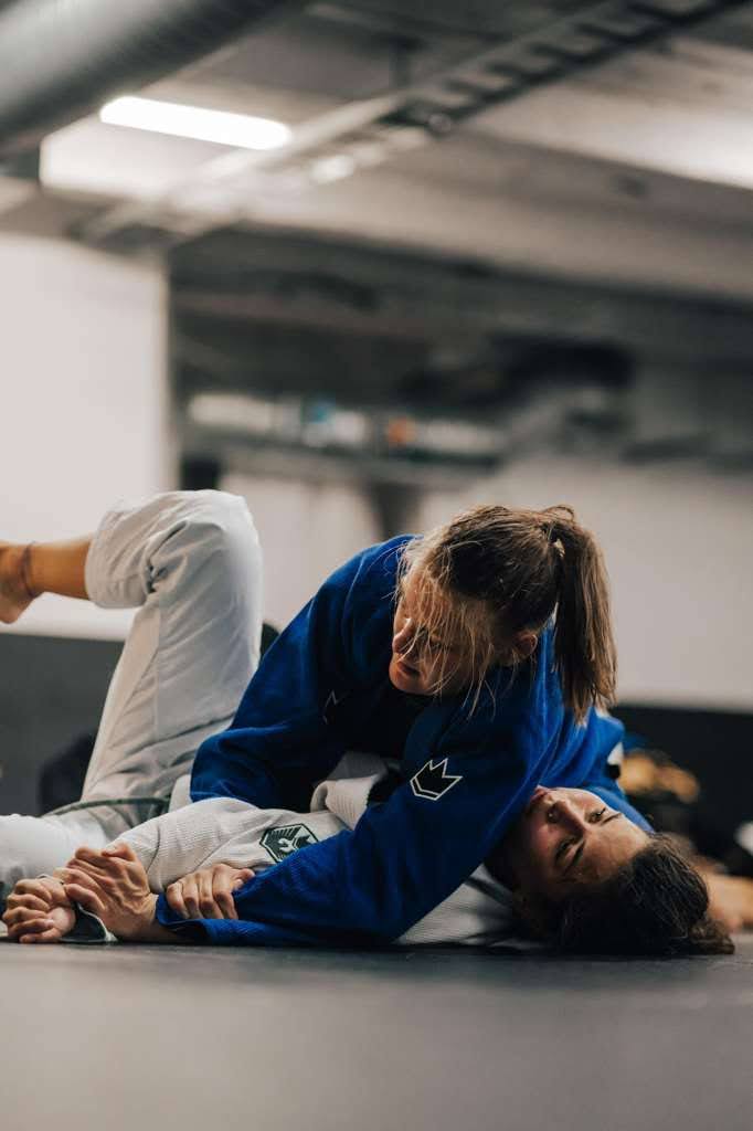 Two women practicing Brazilian Jiu-Jitsu on the mat, one wearing a blue gi applying side control on her training partner in a white gi.