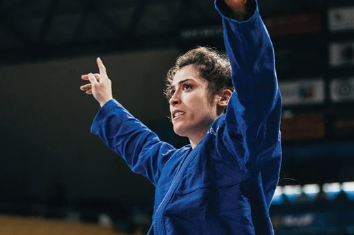 BJJ competitor Abbie O'Toole in blue gi raises arms in victory celebration at competition arena.