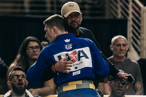 Two people embracing, one wearing a blue BJJ gi with a USA patch, in a crowded indoor setting.