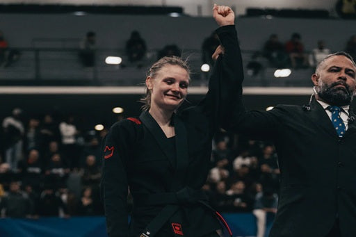 Astrid Scholin celebrates victory in black gi with raised arm at BJJ competition, standing beside official in arena.