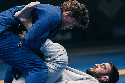 Two martial artists in blue and white gis engage in a ground grappling match on a mat during a Brazilian Jiu-Jitsu competition.