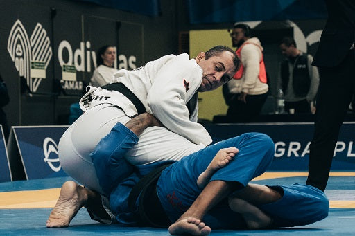 Two Jiu-Jitsu competitors engage on the mat during a match, one wearing a white gi and the other a blue gi.