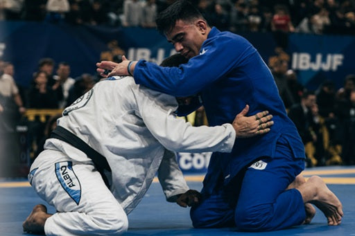 Two grapplers compete in a Brazilian jiu-jitsu match, one in a white gi and the other in blue, engaged in a close hold.