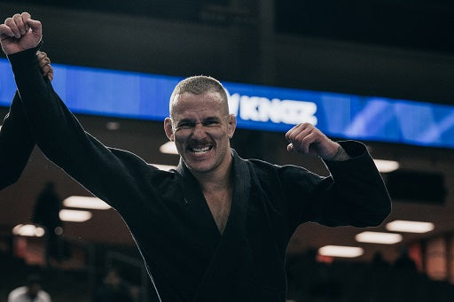A BJJ practitioner in black gi celebrates victory with raised arms inside a sports arena.