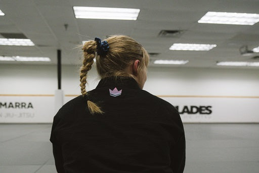 A girl with a braided ponytail sits on a mat, wearing a black jiu-jitsu gi, in a spacious dojo with walls marked by training logos.
