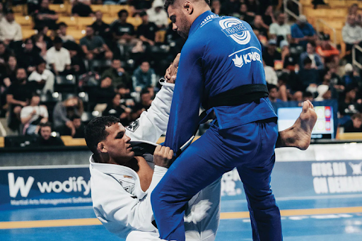 Two men wearing blue and white BJJ uniforms are engaged in a grappling match tournament on the floor.