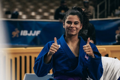 Bia Alves, accomplished Brazilian jiu-jitsu competitor, smiles confidently while giving two thumbs up in blue gi at a tournament event.