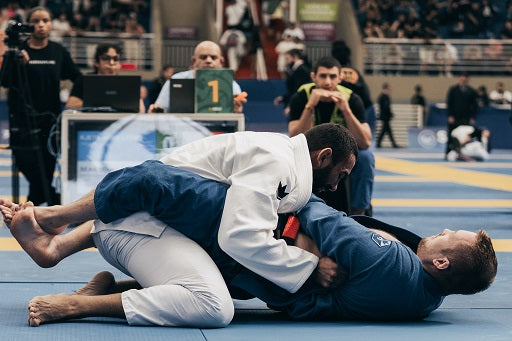 Two BJJ competitors grapple on a blue mat during a tournament match, with a crowd visible in the background.