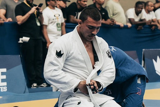 BJJ practitioner Davi Vetoraci de Menezes in a white gi adjusts his belt while standing on the competition mats.