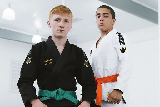 Two young BJJ practitioner wearing gi uniforms, one with a green belt and black gi, the other with an orange belt and white gi, posing indoors.