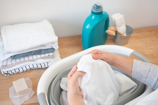 Person carefully hand-washing white gi in basin with liquid detergent and cleaning supplies, demonstrating proper martial arts uniform washing care.