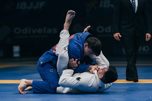 Two male martial artists competing in a Brazilian Jiu-Jitsu match, wearing blue and white gis on a mat with a referee nearby.
