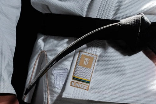 Close-up of a person wearing a Kingz white BJJ gi with a black belt tied around the waist, showing brand patches on the gi.