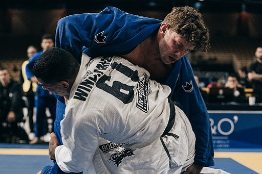 Two men in gi uniforms engaged in a Brazilian Jiu-Jitsu match on a competition mat.