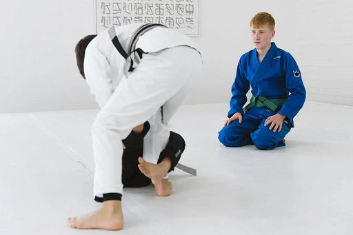 Two kids, one in white and one in blue gi with green belt, practicing Brazilian Jiu-Jitsu on a white mat indoors.