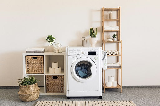 Laundry room with a front-loading washing machine, wooden shelf with towels and toilet paper, wicker baskets, and potted plants.