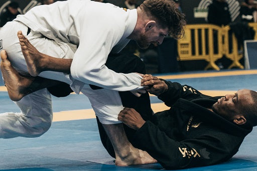 Two martial artists in white and black gis engage in a Brazilian Jiu-Jitsu match on a blue mat with spectators in the background.