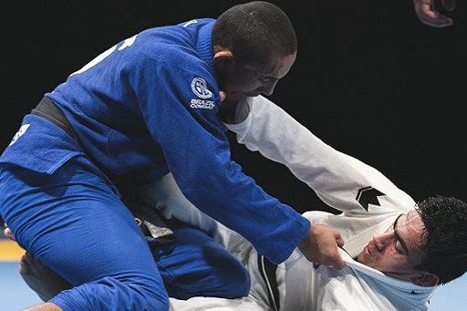 Two martial artists compete in Brazilian Jiu-Jitsu, one wearing a blue gi and the other a white gi, on a mat.