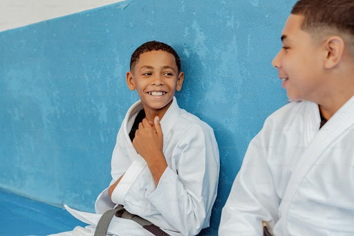 Two children in white gis sitting and resting against a blue wall in a BJJ training area.