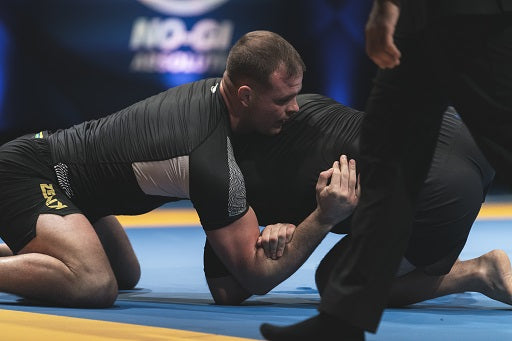 Two men grappling in a Brazilian Jiu-Jitsu no-gi match on a blue mat, with a referee observing closely.