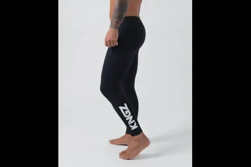 Side view of a shirtless person wearing black spats with KNGZ printed near the ankle, standing barefoot against a plain background.