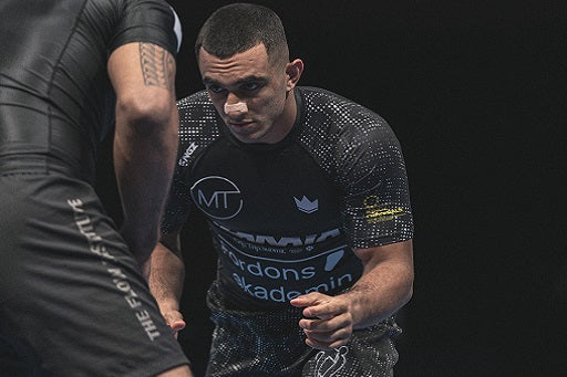 Two BJJ athletes in black rash guard face each other in a wrestling or grappling stance against a dark background.